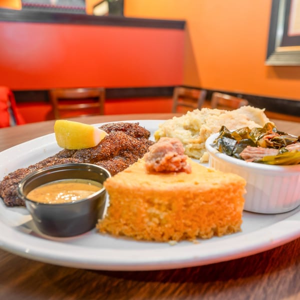 Southern meal with blackened fish, cornbread, mashed potatoes, collard greens, and dipping sauce.
