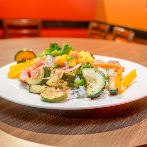 A white plate of grilled zucchini, bell peppers, and broccoli over rice on a wooden table.